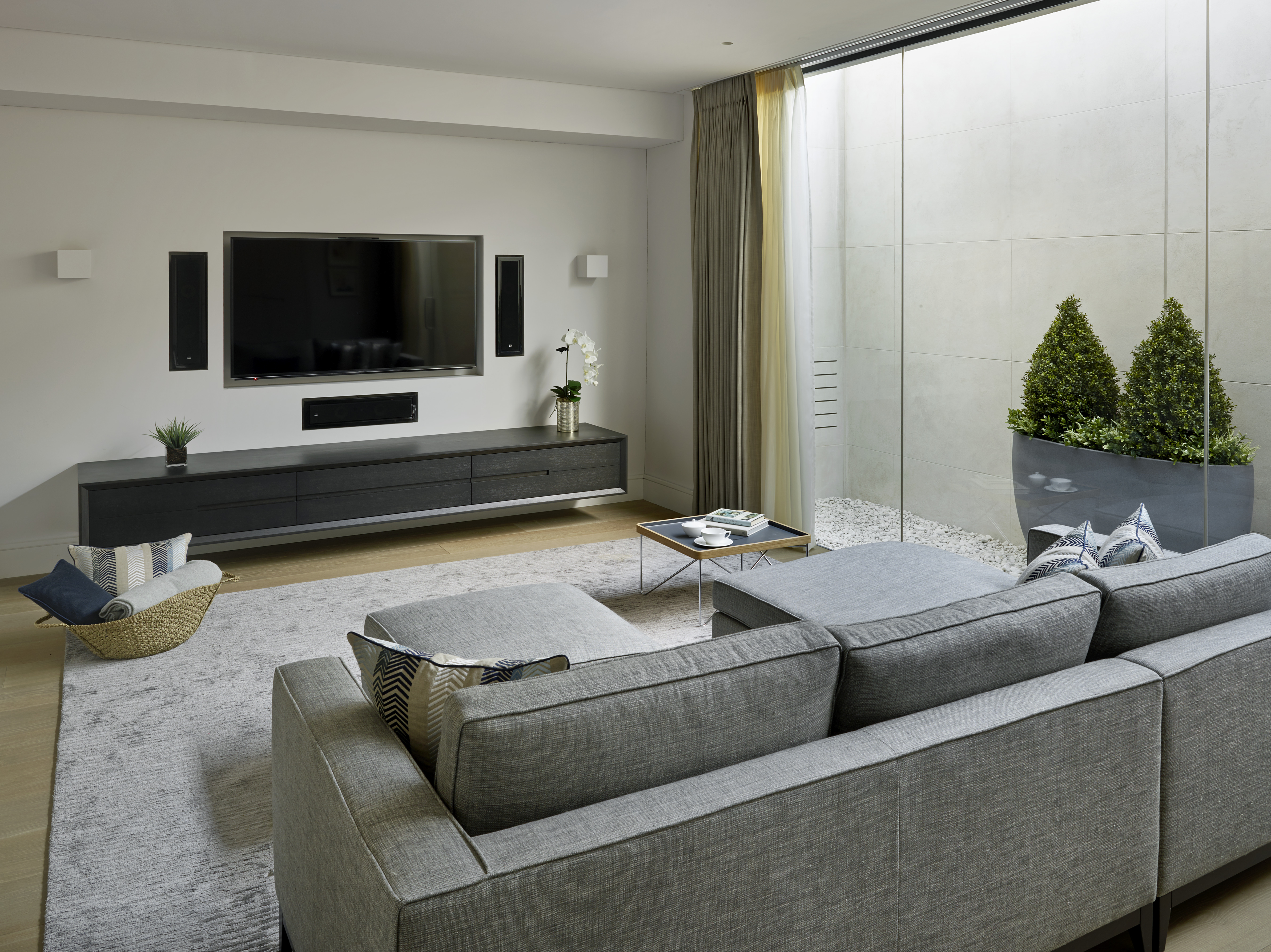 Contemporary movie room in basement with full length glass wall leading to stone clad basement lightwell with clipped hedge planters. 