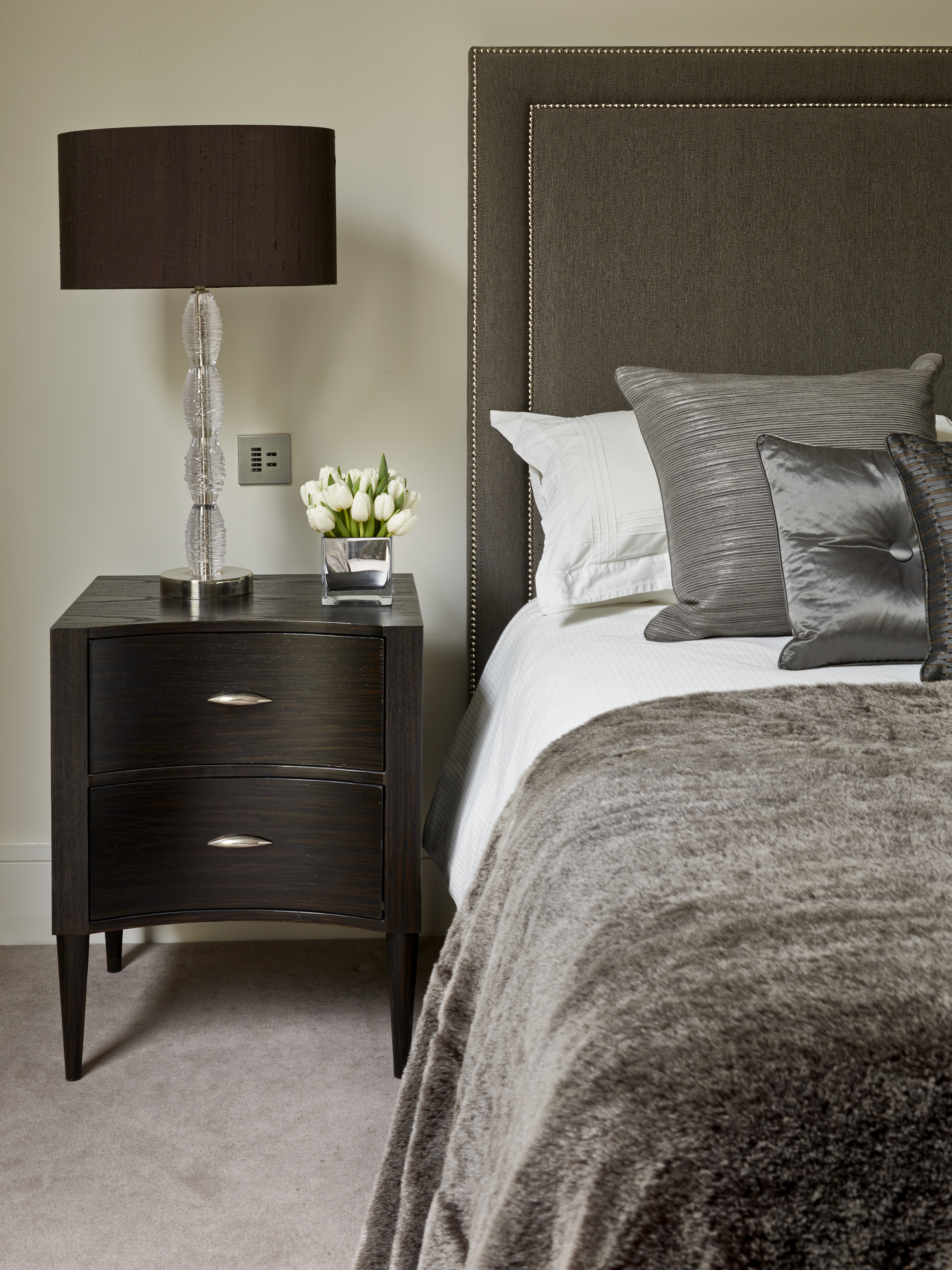 Classically styled upholstered headboard with studded border, decorative cushions and fur throw alongside curved dark oak bedside table.